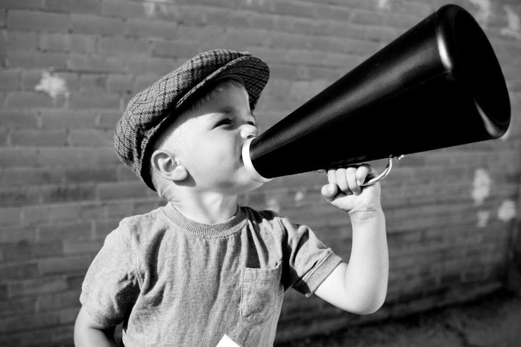 Young child wearing a flat cap and t-shirt holds a megaphone to their mouth, appearing to shout or speak loudly. The background is an out-of-focus brick wall. The image is in black and white. | sjhstudios