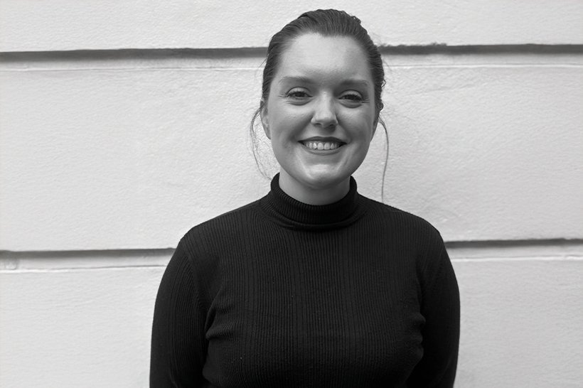 A woman with dark hair pulled back, wearing a black turtleneck sweater, stands smiling in front of a light-colored, textured wall. The photo is in black and white. | sjhstudios