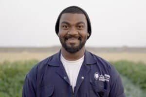 A man wearing a dark beanie and a blue work jacket stands outdoors, smiling at the camera with green fields blurred in the background. | sjhstudios