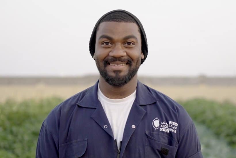A man wearing a dark beanie and a blue work jacket stands outdoors, smiling at the camera with green fields blurred in the background. | sjhstudios