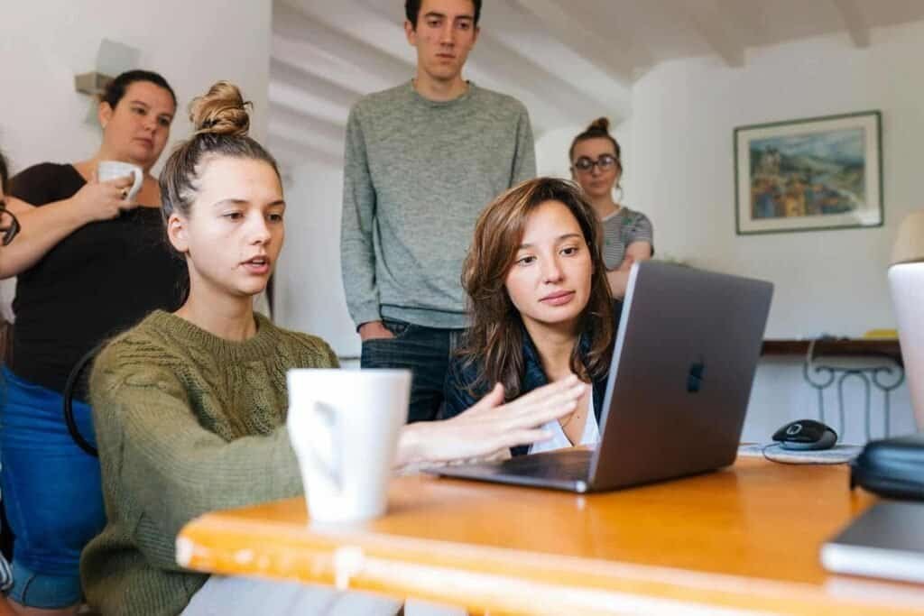 Five people are gathered around a table, focused on a laptop. Two women sit at the table, one gesturing toward the screen, while three others stand behind them, observing. Coffee mugs and a mouse are also on the table. | sjhstudios