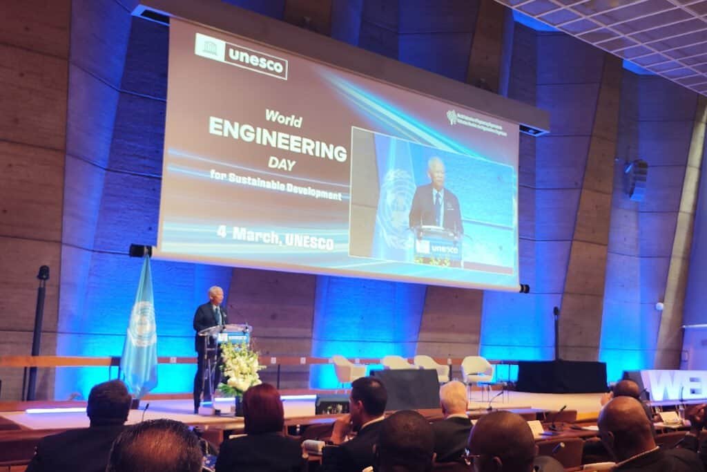 A speaker stands at a podium on stage during World Engineering Day for Sustainable Development at UNESCO, with a large screen displaying event details and an audience seated in the foreground. | sjhstudios