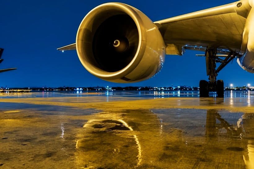 A close-up view of a jet engine and wing of an airplane on a wet airport tarmac at night, with city lights and their reflections visible in the background. | sjhstudios