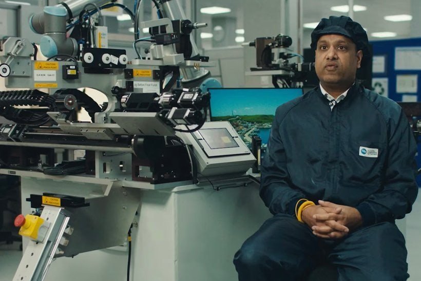 A man in a dark cleanroom suit and cap sits in a lab with advanced machinery and computers behind him, suggesting a high-tech or manufacturing environment. | sjhstudios
