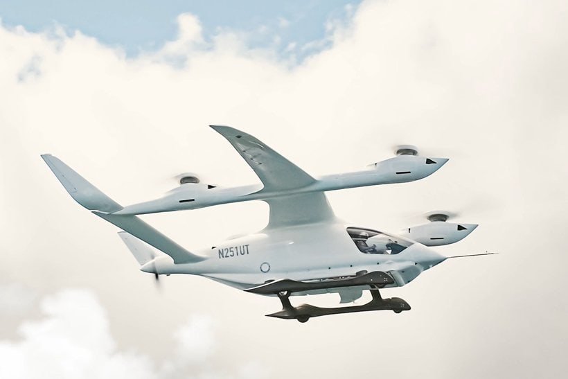 A white electric vertical takeoff and landing (eVTOL) aircraft with four large rotors is flying in the sky against a background of clouds. The aircraft’s registration number, N251UT, is visible on the side. | sjhstudios