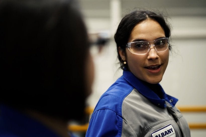 A woman wearing safety glasses and a blue-gray work uniform with a name badge is looking at a colleague in an industrial or laboratory setting. | sjhstudios