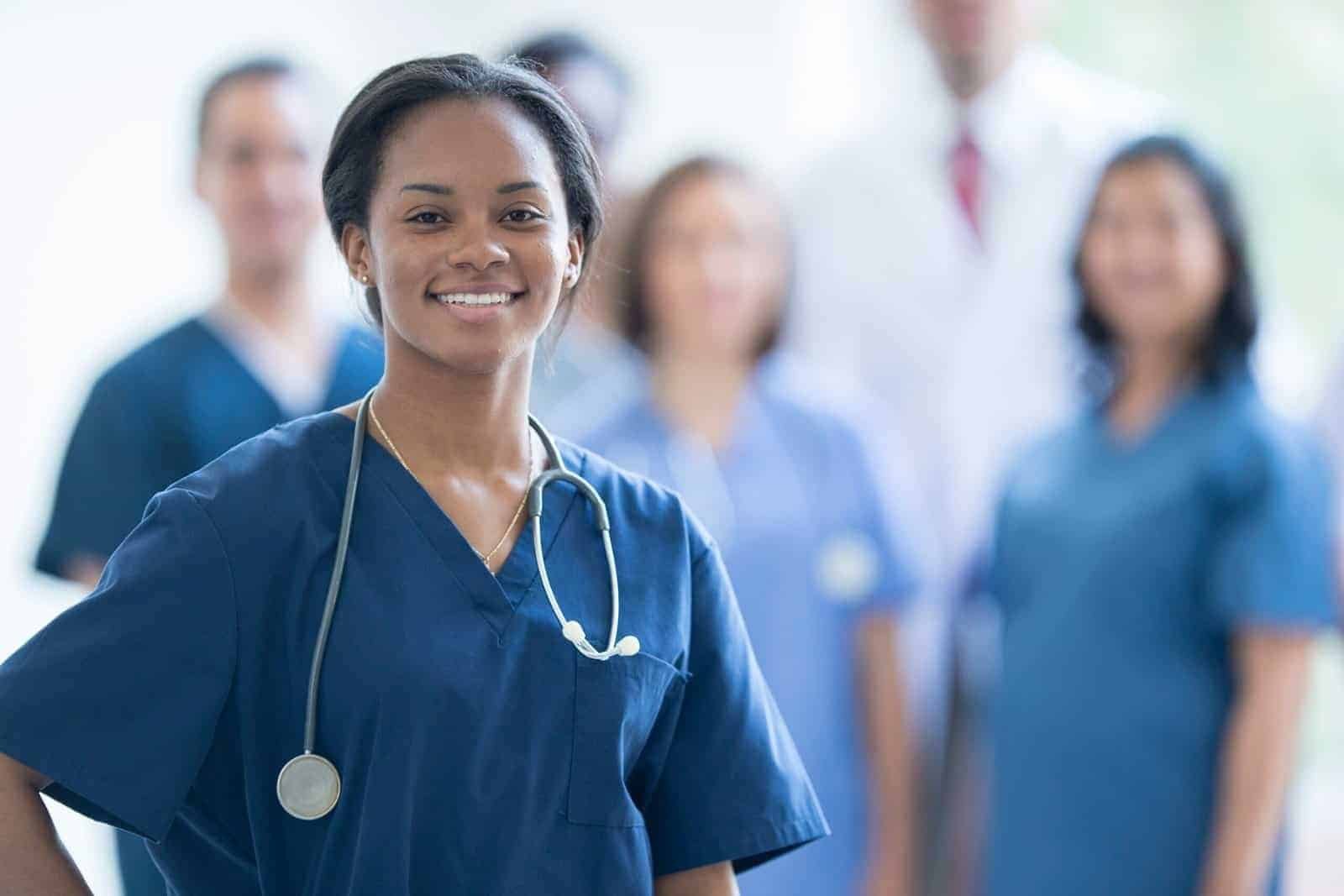 A smiling healthcare worker wearing blue scrubs and a stethoscope stands in the foreground, with other medical professionals in similar attire blurred in the background. | sjhstudios