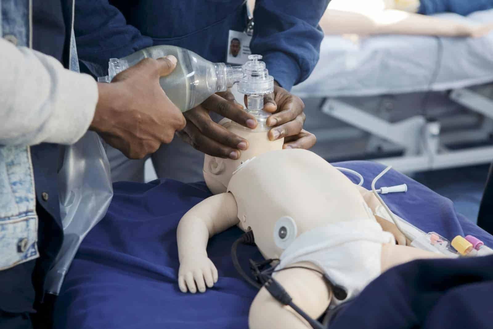 Two people practice emergency medical procedures on an infant mannequin, using a manual resuscitator mask on the mannequin’s face. Medical equipment is visible on the table. | sjhstudios