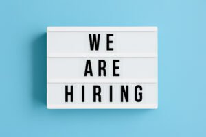 A white lightbox sign with black letters displays the message "WE ARE HIRING" against a light blue background. | sjhstudios